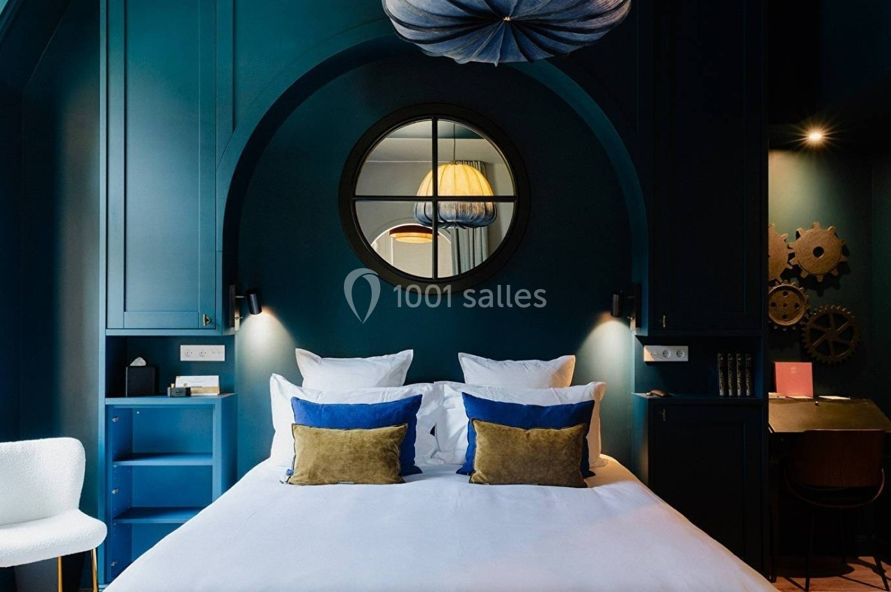 Chambre moderne avec lit double, coussins colorés, mur bleu, miroir rond et bureau éclairé dans un coin.