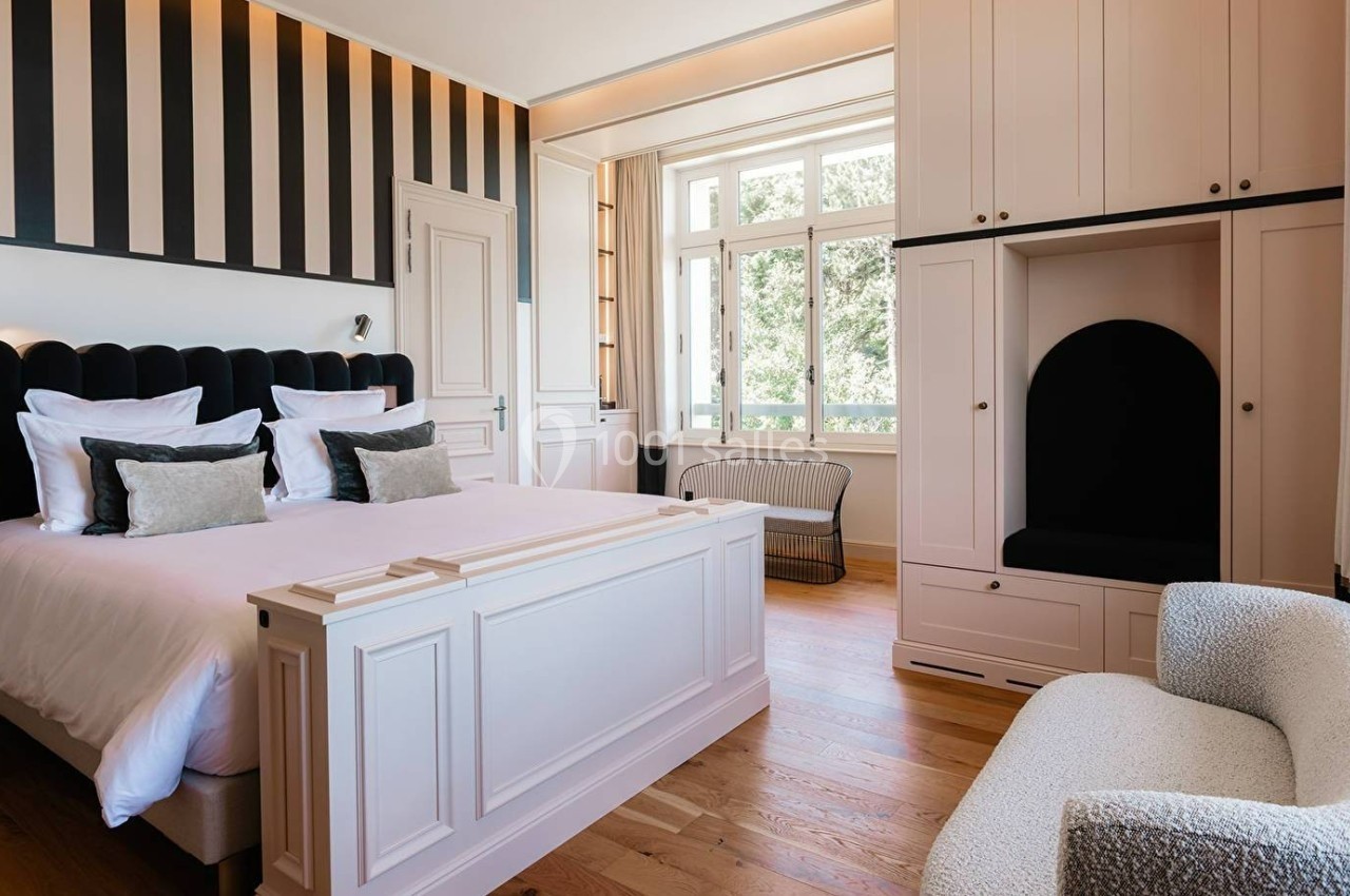 Chambre lumineuse avec lit double, mobilier blanc, fauteuil texturé et grande fenêtre donnant sur un espace verdoyant.