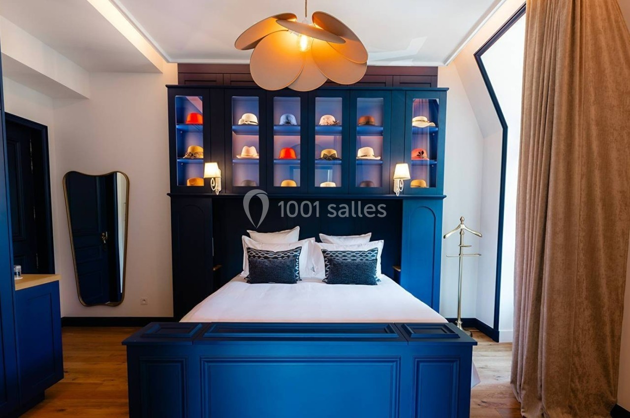 Chambre avec lit double, mobilier bleu, étagères décorées de chapeaux, éclairage chaleureux et rideaux beiges.