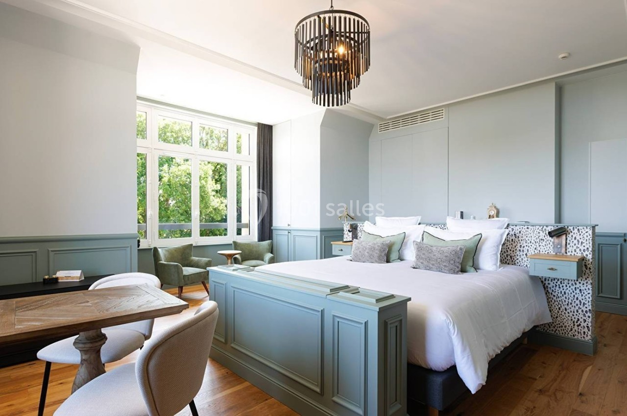 Chambre lumineuse avec lit double, mobilier en bois clair, fauteuils verts et grande fenêtre donnant sur un espace arboré.