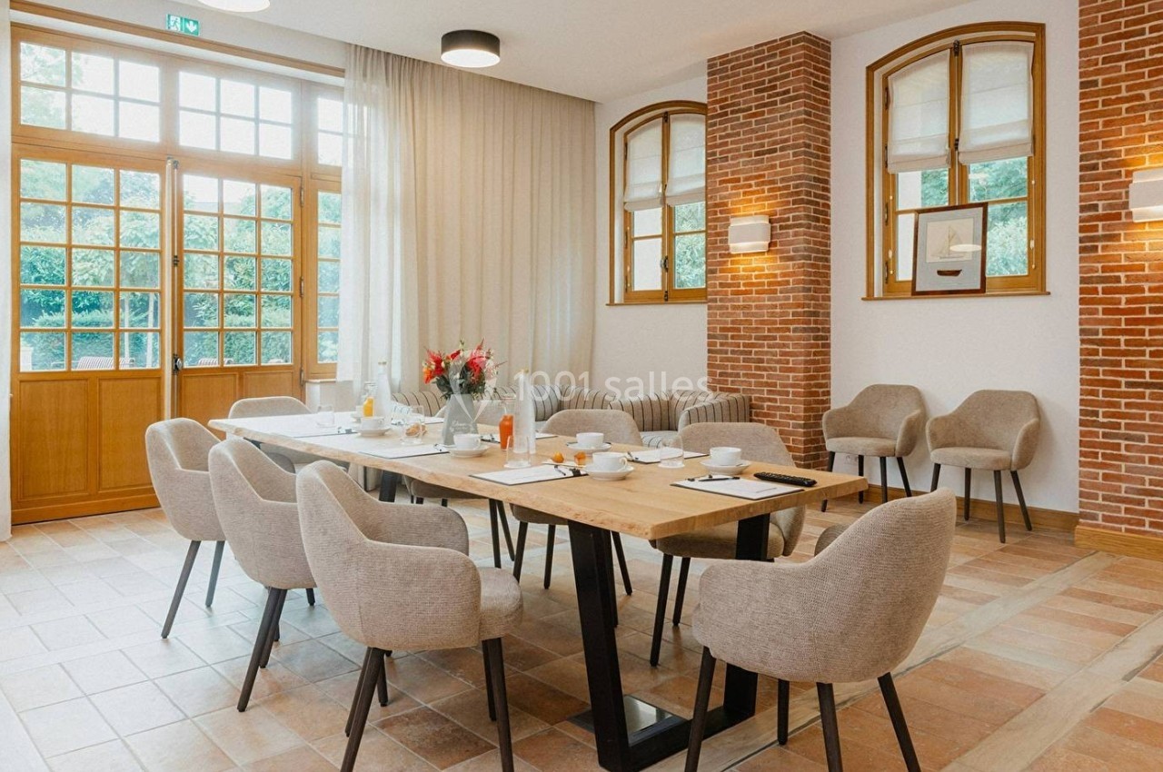 Salle lumineuse avec table en bois dressée pour un repas, entourée de chaises beige et murs en briques apparentes.