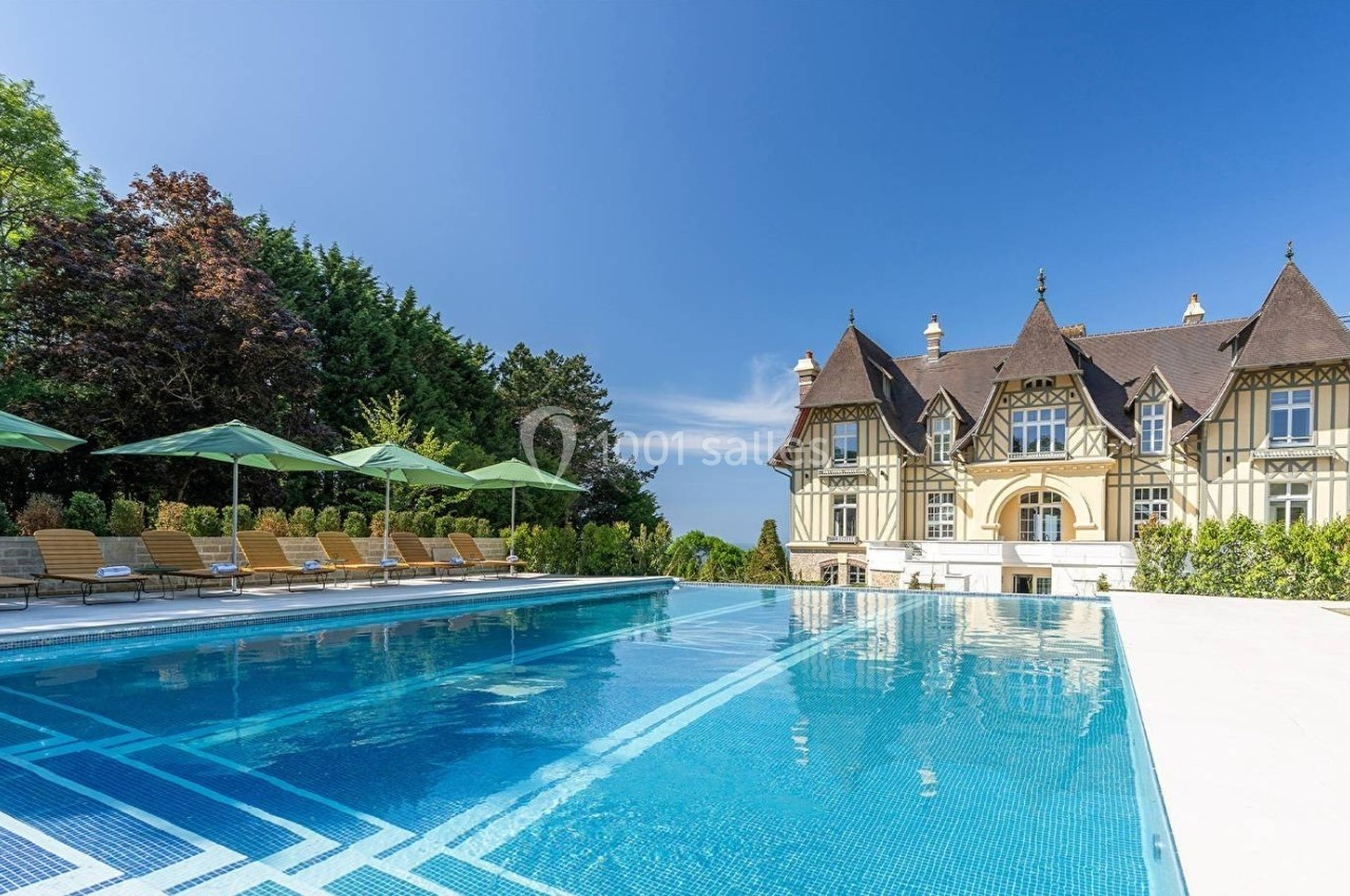 Piscine extérieure avec transats et parasols, devant un manoir de style traditionnel entouré de verdure.