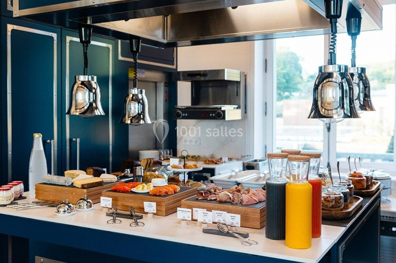 Buffet de petit-déjeuner avec boissons, charcuterie, fromages, viennoiseries et condiments sur un comptoir moderne.