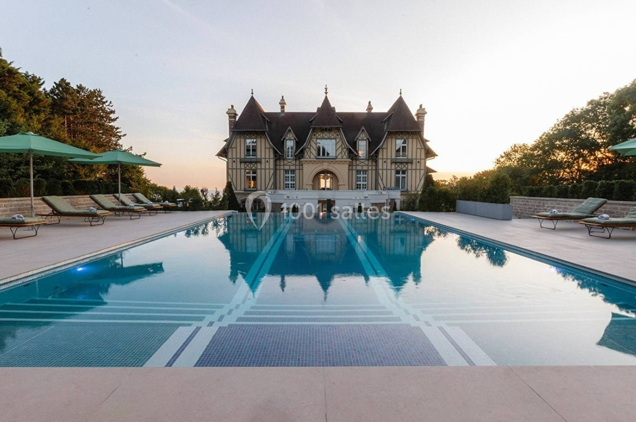 Piscine extérieure avec transats et parasols, devant un manoir à colombages entouré de verdure au coucher du soleil.