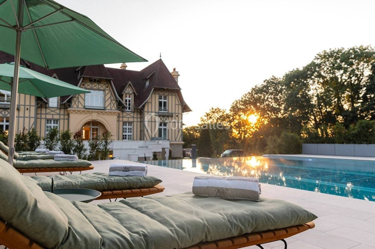 Chaises longues avec coussins verts près d'une piscine, devant un manoir à colombages au coucher du soleil.