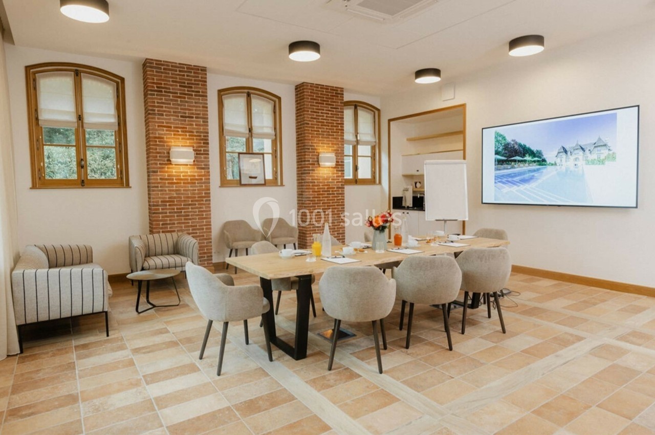 Salle de réunion lumineuse avec table en bois, chaises beiges, écran mural et murs en briques apparentes.