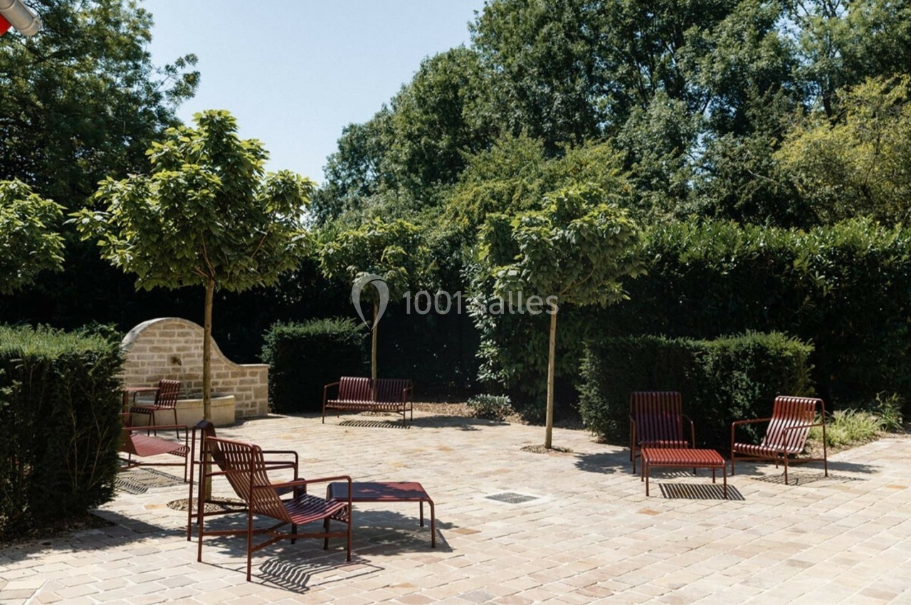 Cour pavée avec des chaises longues rouges, entourée de haies et d'arbres sous un ciel dégagé.