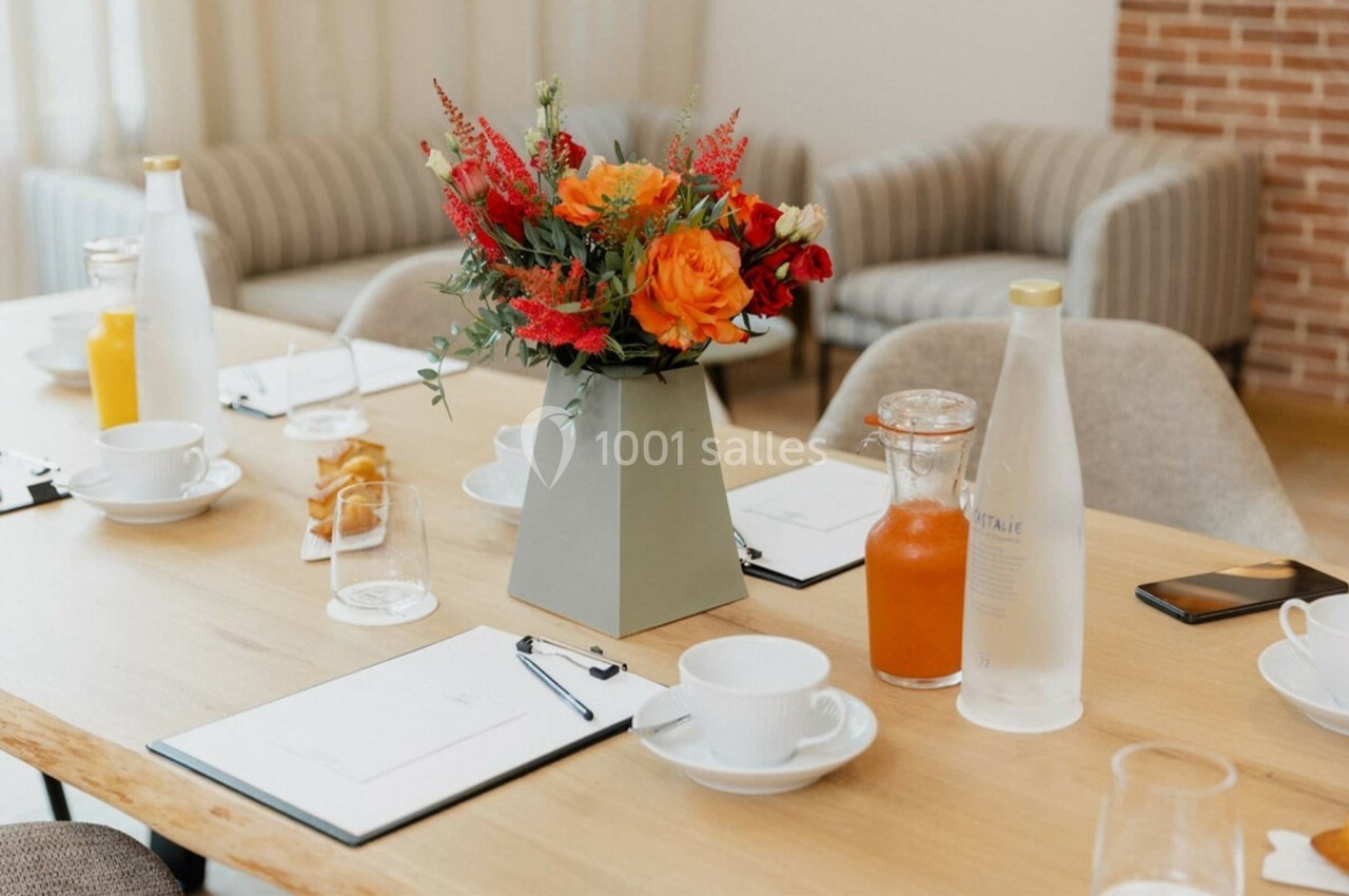 Vase de fleurs colorées sur une table en bois dressée avec des documents, tasses, carafes et jus d'orange.