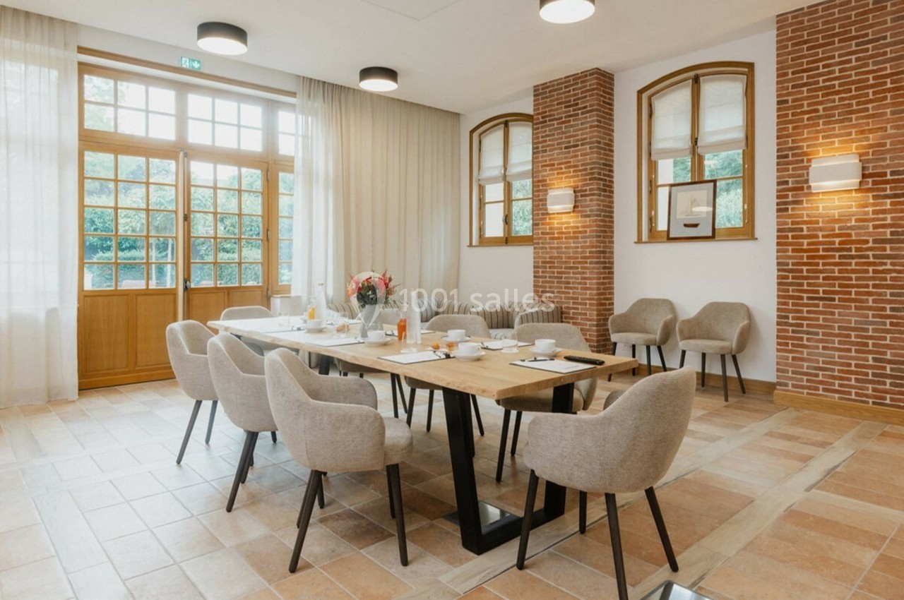 Salle lumineuse avec table en bois, chaises beige, grandes fenêtres, murs en briques et vaisselle disposée.