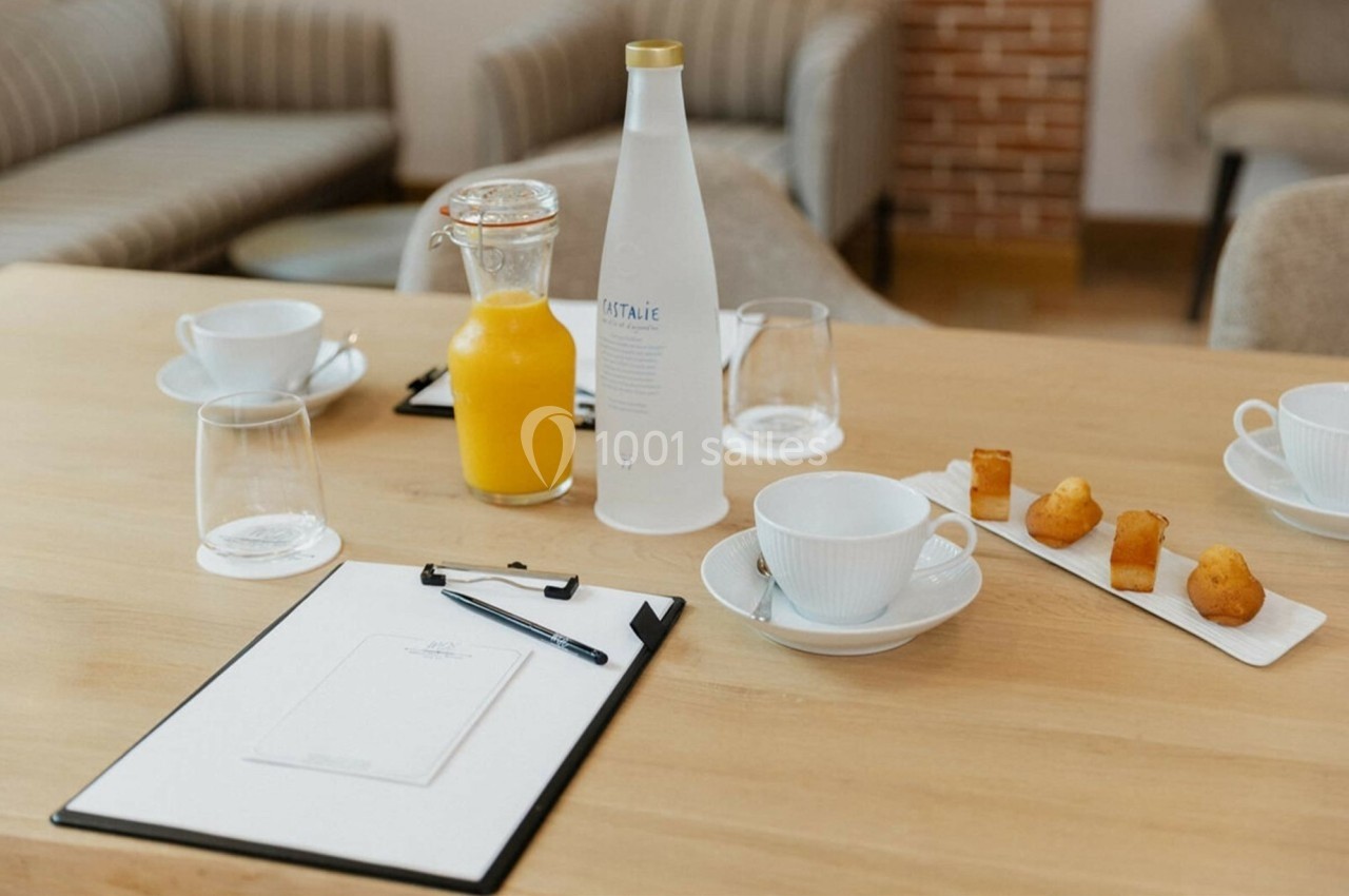 Table en bois avec carafe de jus d'orange, bouteille d'eau, tasses, verres, bloc-notes et assiette de petits gâteaux.