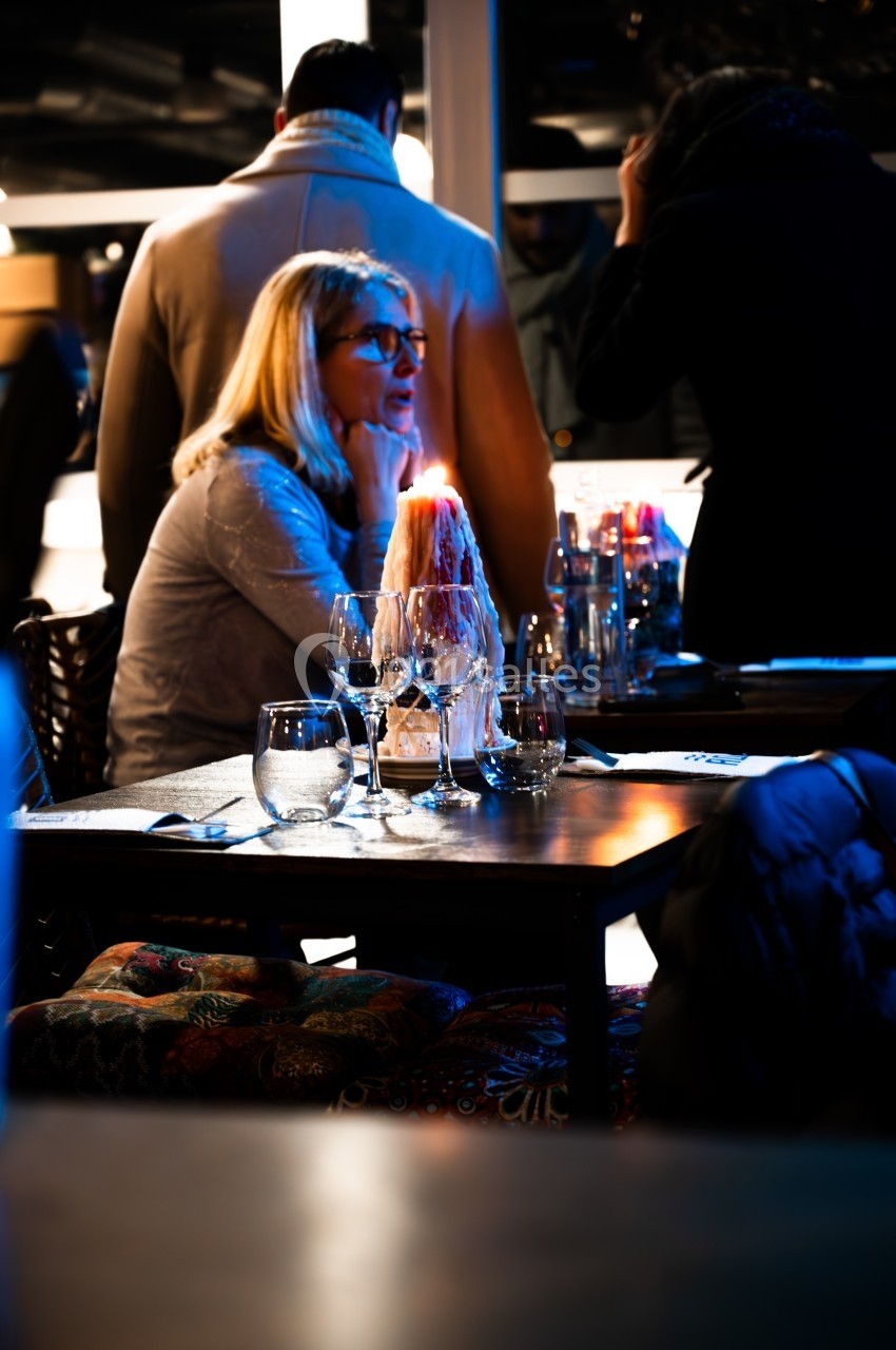 Une femme assise à une table de restaurant éclairée par une bougie, entourée de convives en arrière-plan flou.