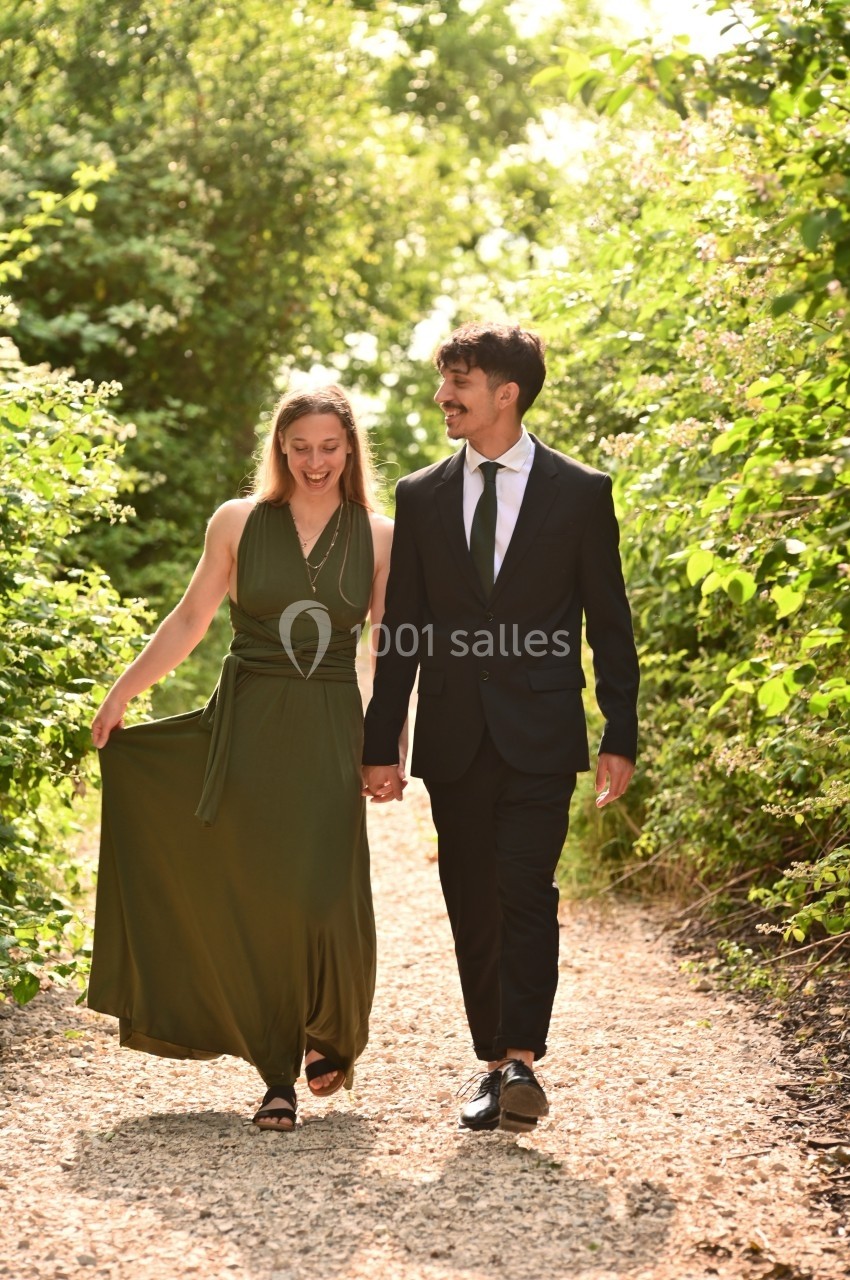Un couple souriant marche main dans la main sur un chemin bordé de verdure par une journée ensoleillée.