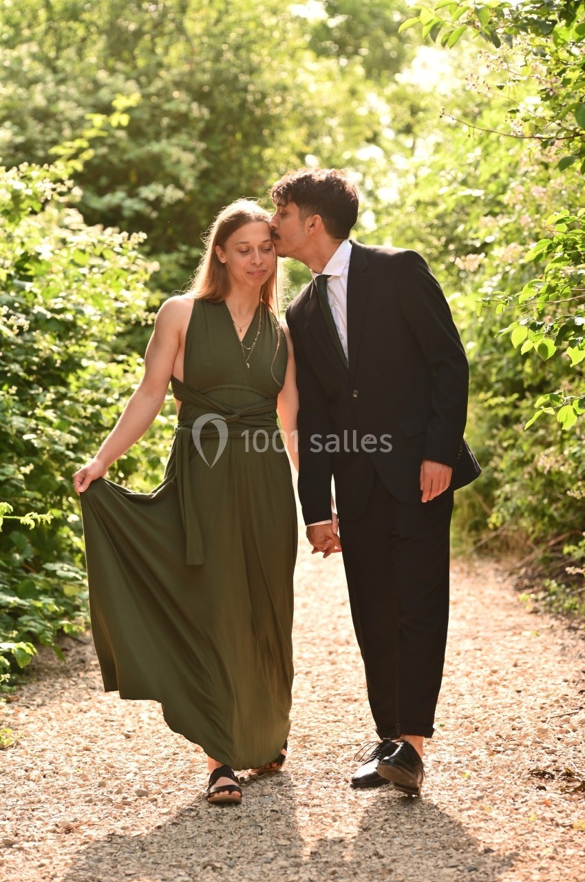 Un couple élégant marche main dans la main sur un chemin bordé de verdure, sous une lumière douce.