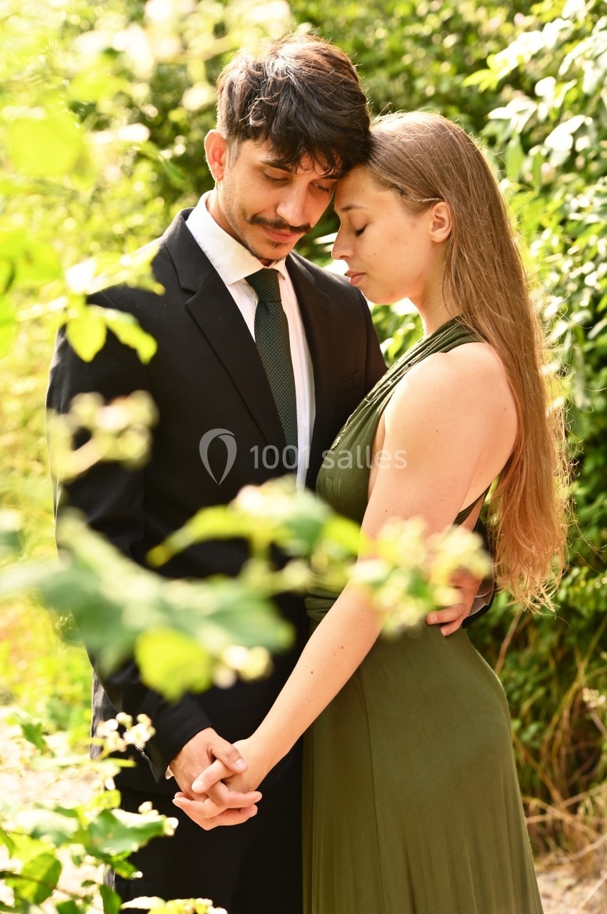 Un couple élégant, l'homme en costume et la femme en robe verte, se tient tendrement dans un jardin verdoyant.