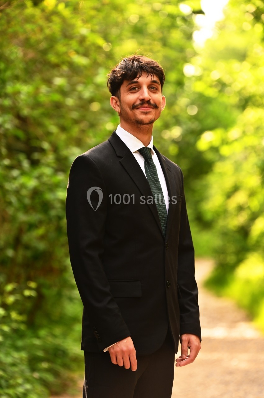 Un homme en costume noir et cravate verte se tient debout sur un chemin bordé de verdure.