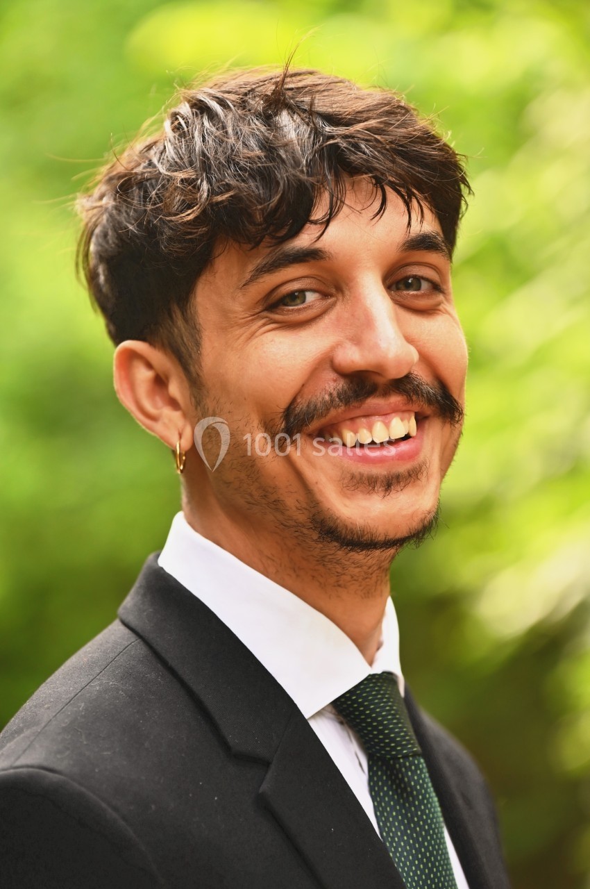 Homme souriant en costume noir et cravate verte, photographié en extérieur avec un fond flou de verdure.