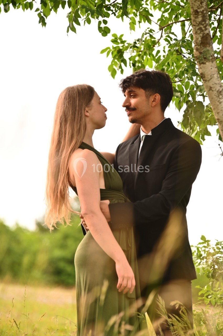 Un couple debout dans un champ verdoyant, échangeant un regard tendre sous un arbre.