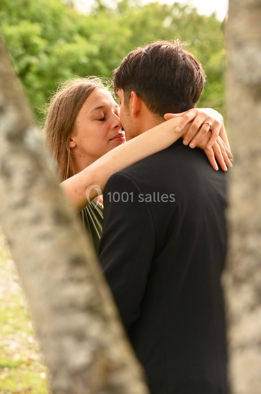 Une femme enlace un homme en costume dans un cadre extérieur verdoyant, vue partiellement encadrée par des arbres.