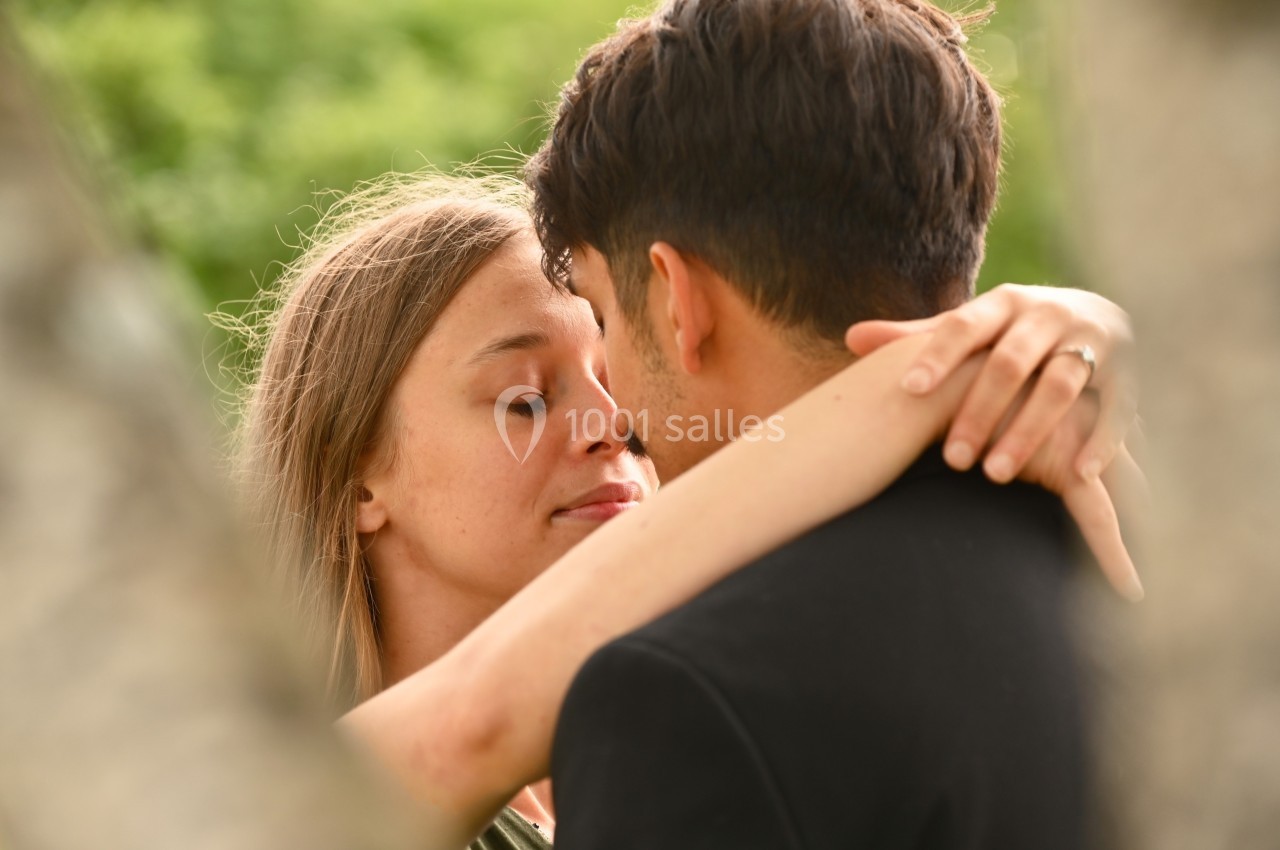Un couple s'enlace tendrement dans un cadre extérieur verdoyant, capturé entre des branches floues au premier plan.
