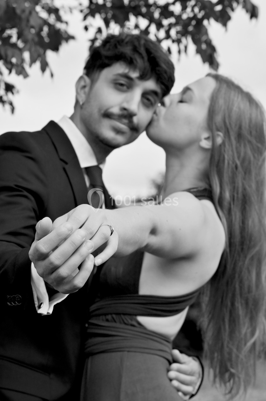 Un couple élégant pose en extérieur, la femme embrassant l'homme sur la joue tout en tenant sa main.