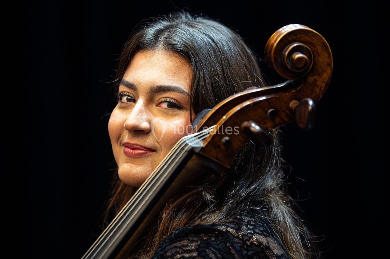 Une femme souriante pose avec un violoncelle, éclairée par une lumière douce sur fond noir.