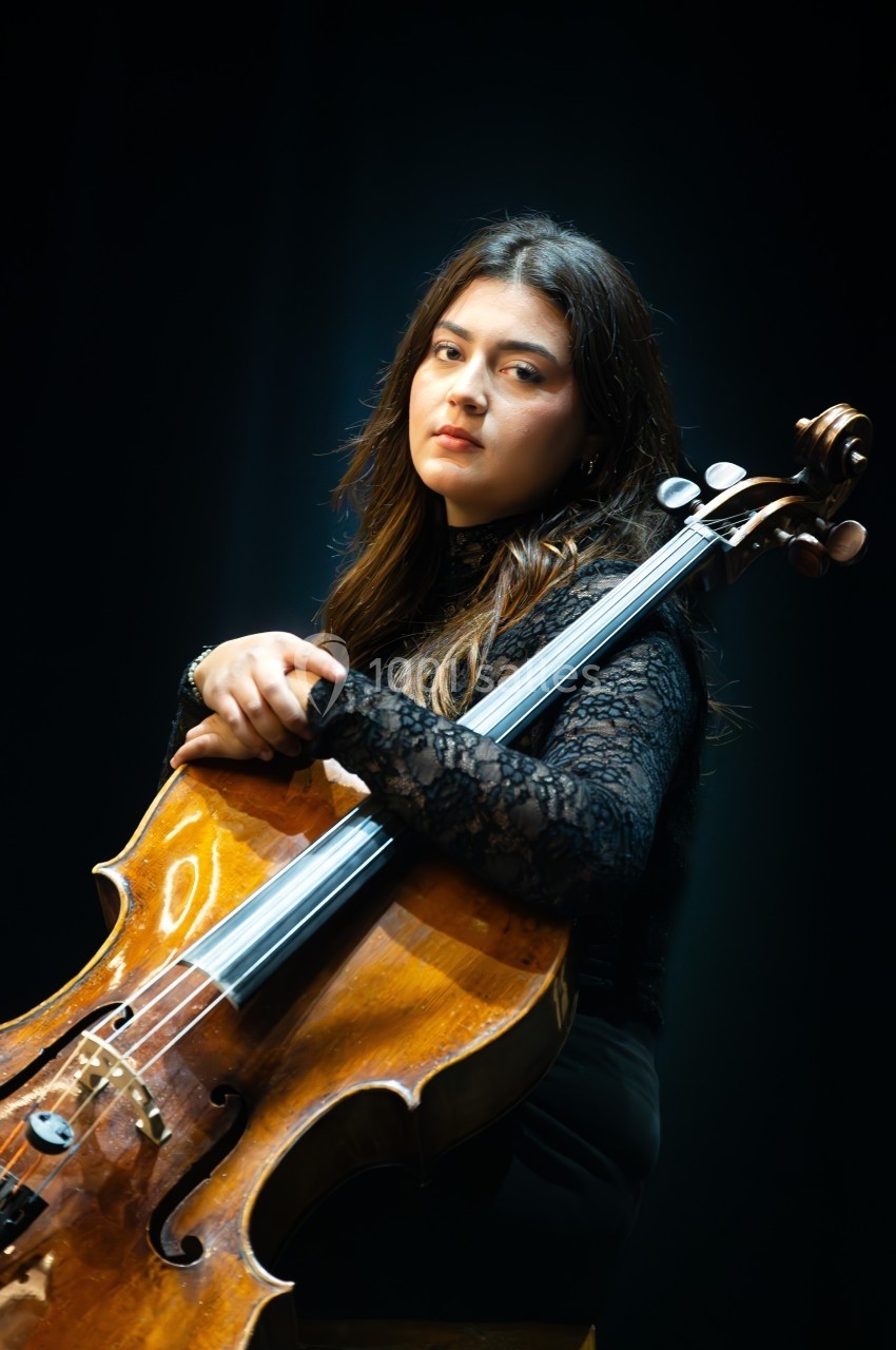 Une femme assise tenant un violoncelle, vêtue de noir, éclairée par une lumière douce sur un fond sombre.