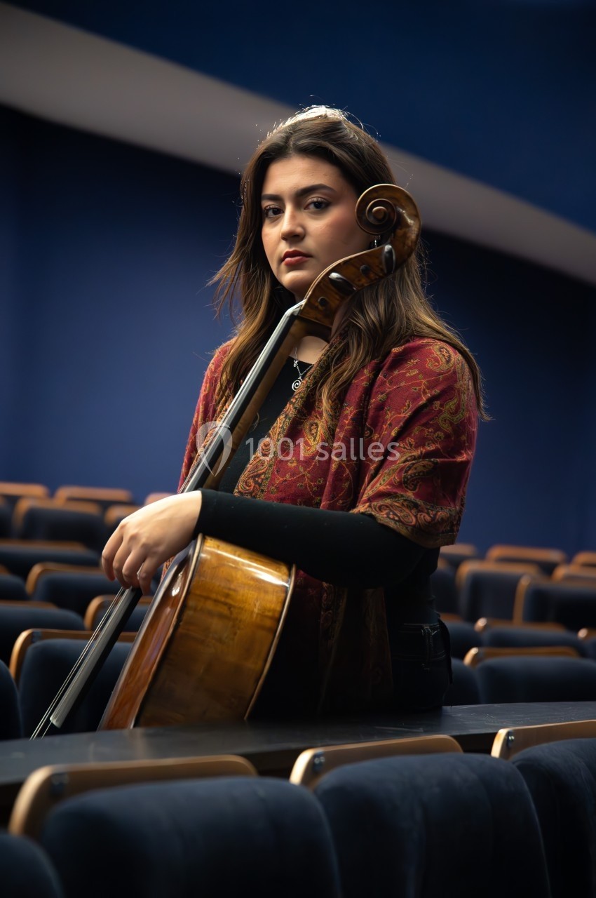 Une femme assise dans une salle vide tient un violoncelle, vêtue d'un châle rouge à motifs.