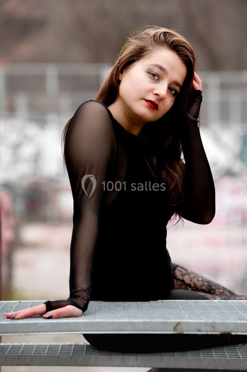 Femme en tenue noire assise sur une structure métallique, regardant l'objectif avec un fond flou urbain.