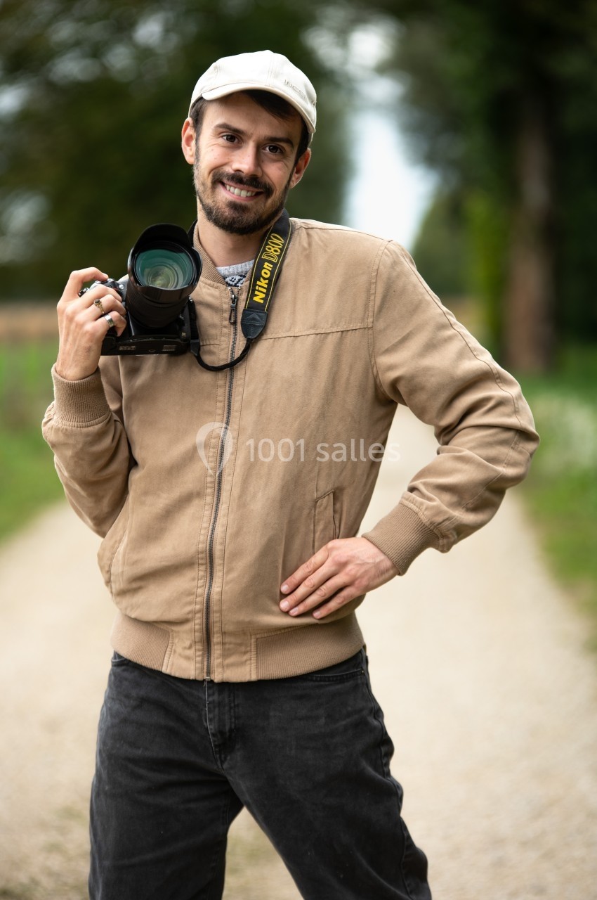 Un homme souriant tenant un appareil photo sur un chemin bordé d'arbres.