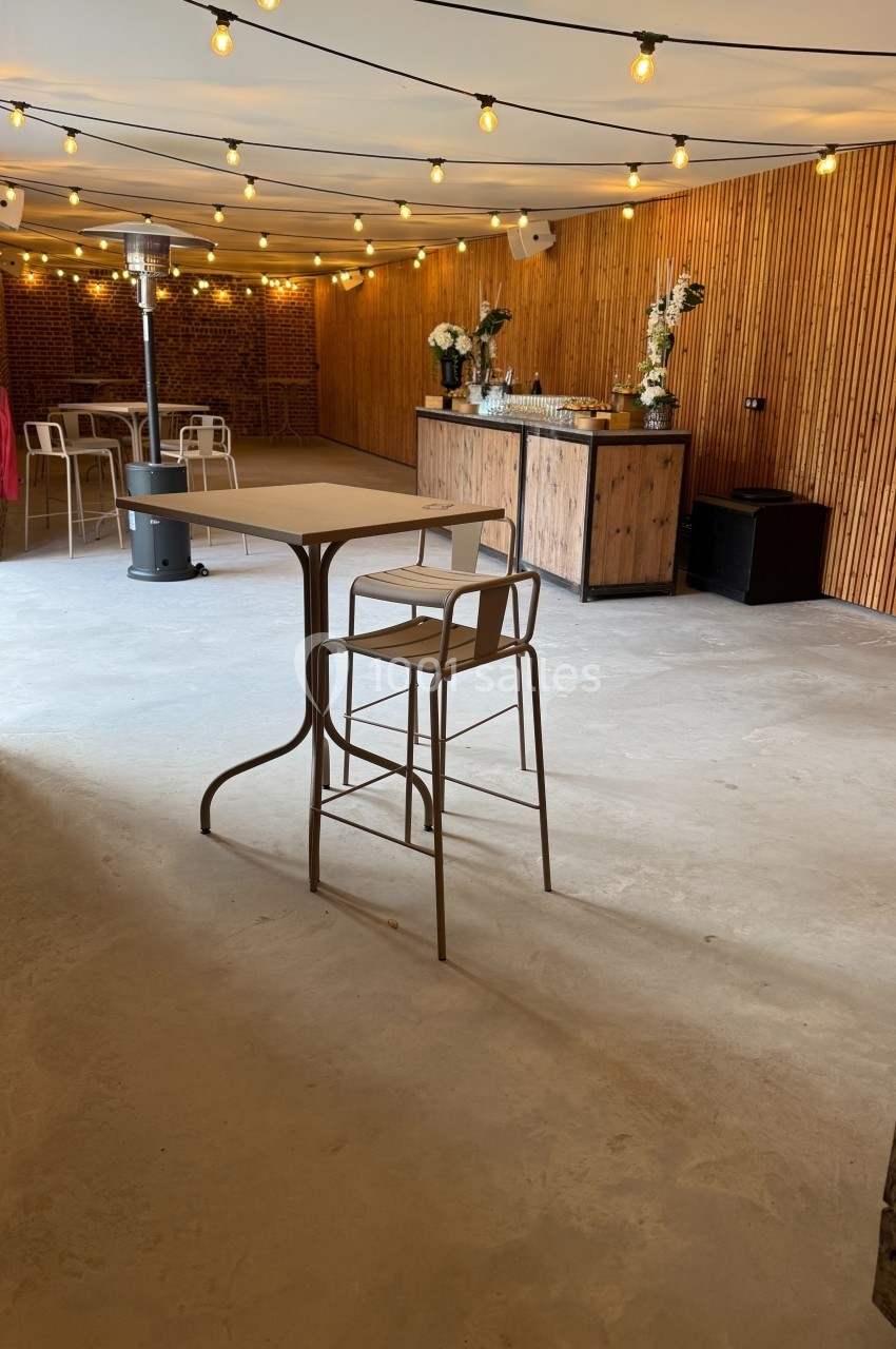 Salle lumineuse avec murs en bois, guirlandes lumineuses, bar en bois, tables hautes et chaises.