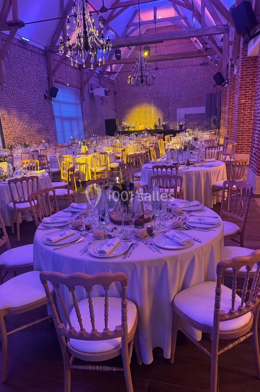 Salle de réception décorée pour un dîner, avec tables rondes dressées, chaises en bois et éclairage tamisé.