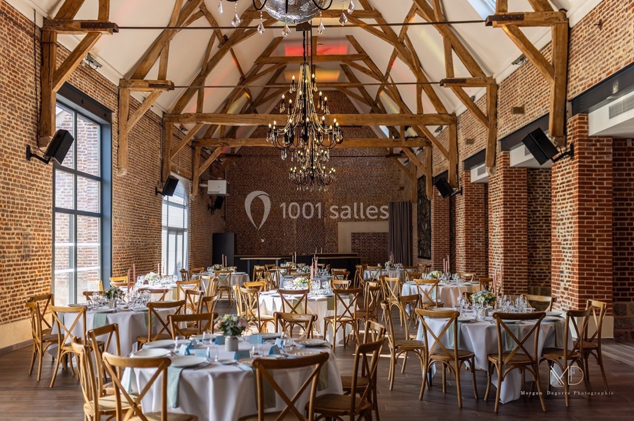 Salle de réception avec poutres apparentes, murs en briques, tables rondes dressées et lustre central élégant.