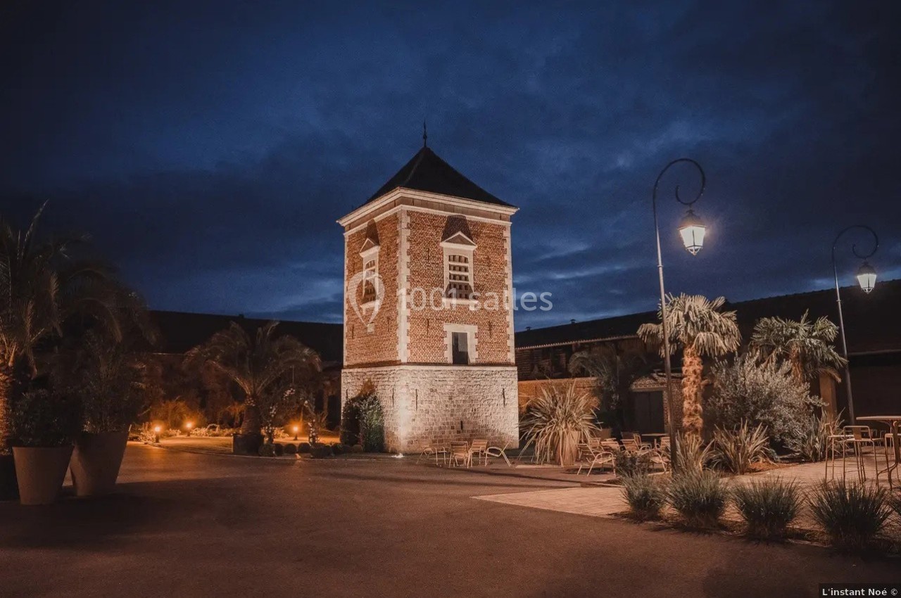 Tour en briques éclairée la nuit, entourée de palmiers et de lampadaires dans une cour pavée.