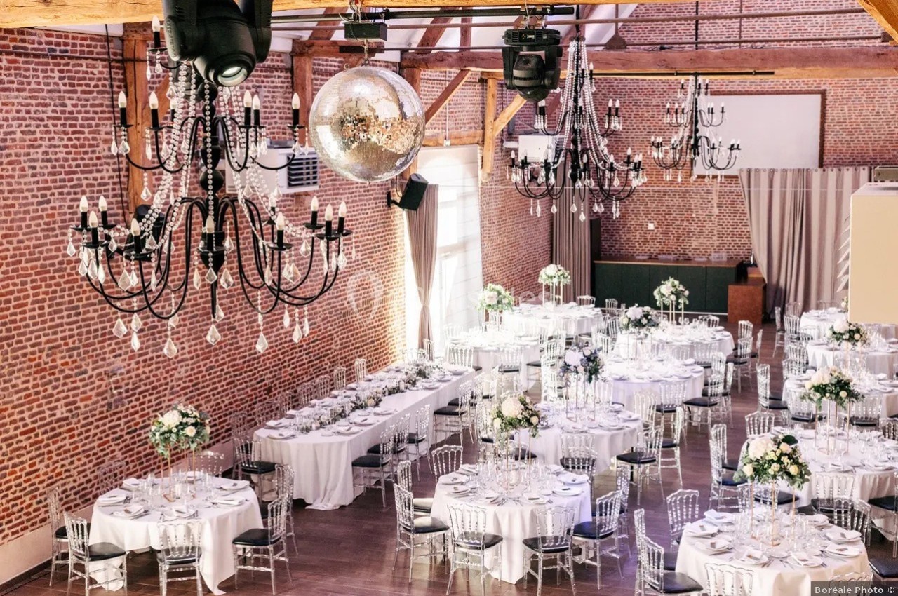 Salle de réception décorée avec des tables rondes dressées, des chandeliers noirs suspendus et une boule à facettes.