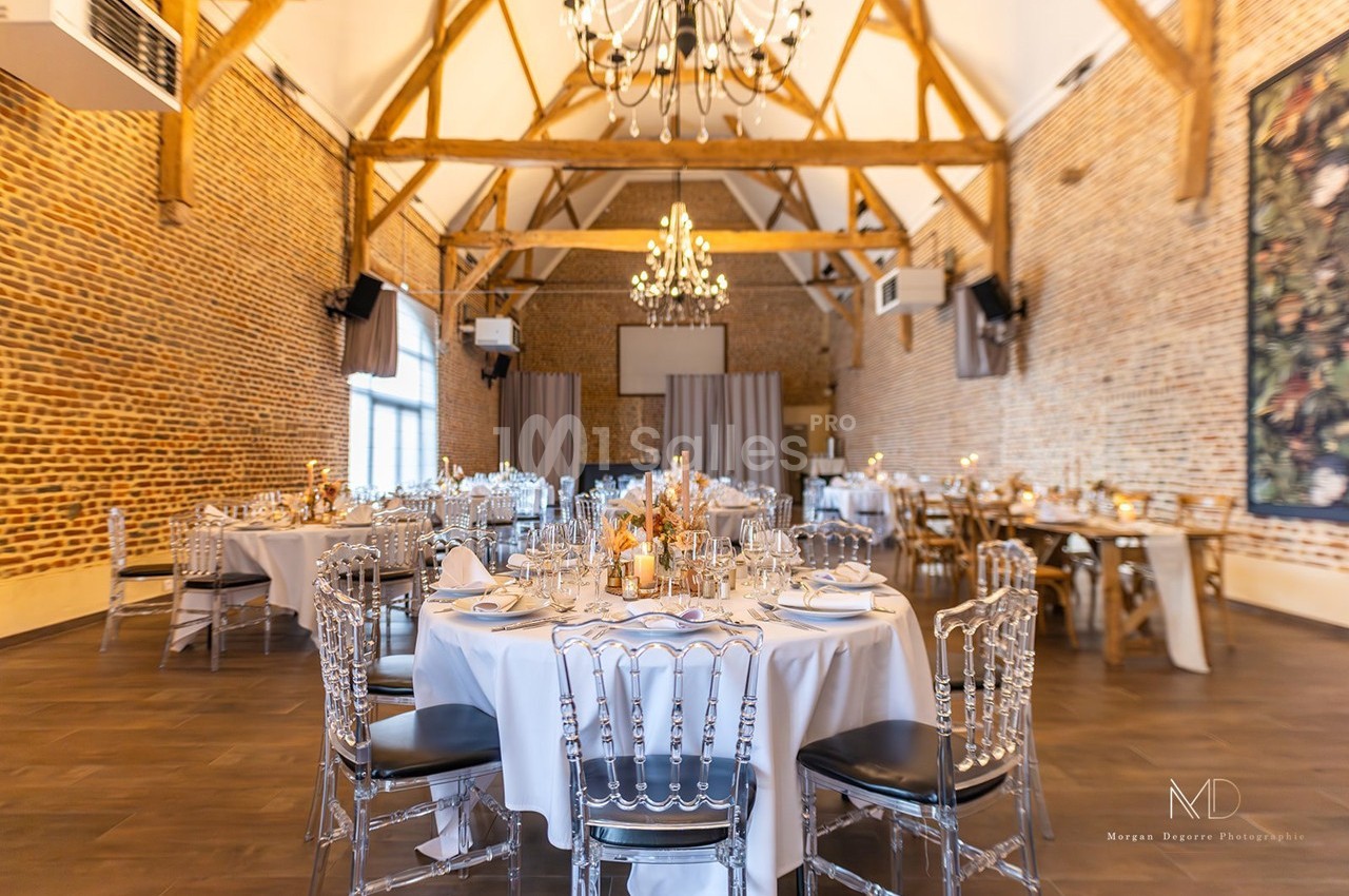Salle de réception lumineuse avec murs en briques, poutres apparentes, tables rondes dressées et lustres suspendus.