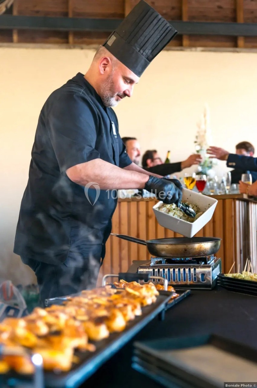 Un chef en tenue noire prépare un plat chaud sur une plaque de cuisson lors d'un événement avec des convives en arrière-plan.