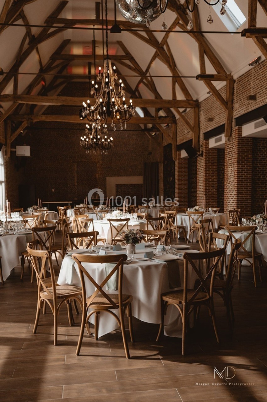Salle de réception lumineuse avec tables rondes dressées, chaises en bois et poutres apparentes au plafond.