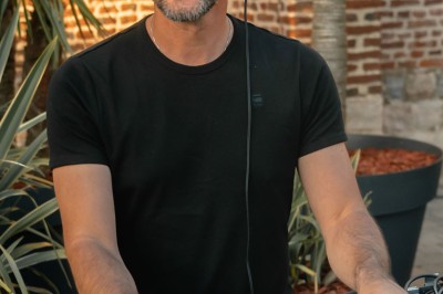 Un DJ mixe en extérieur, portant un casque audio et des lunettes de soleil, avec un mur de briques en arrière-plan.