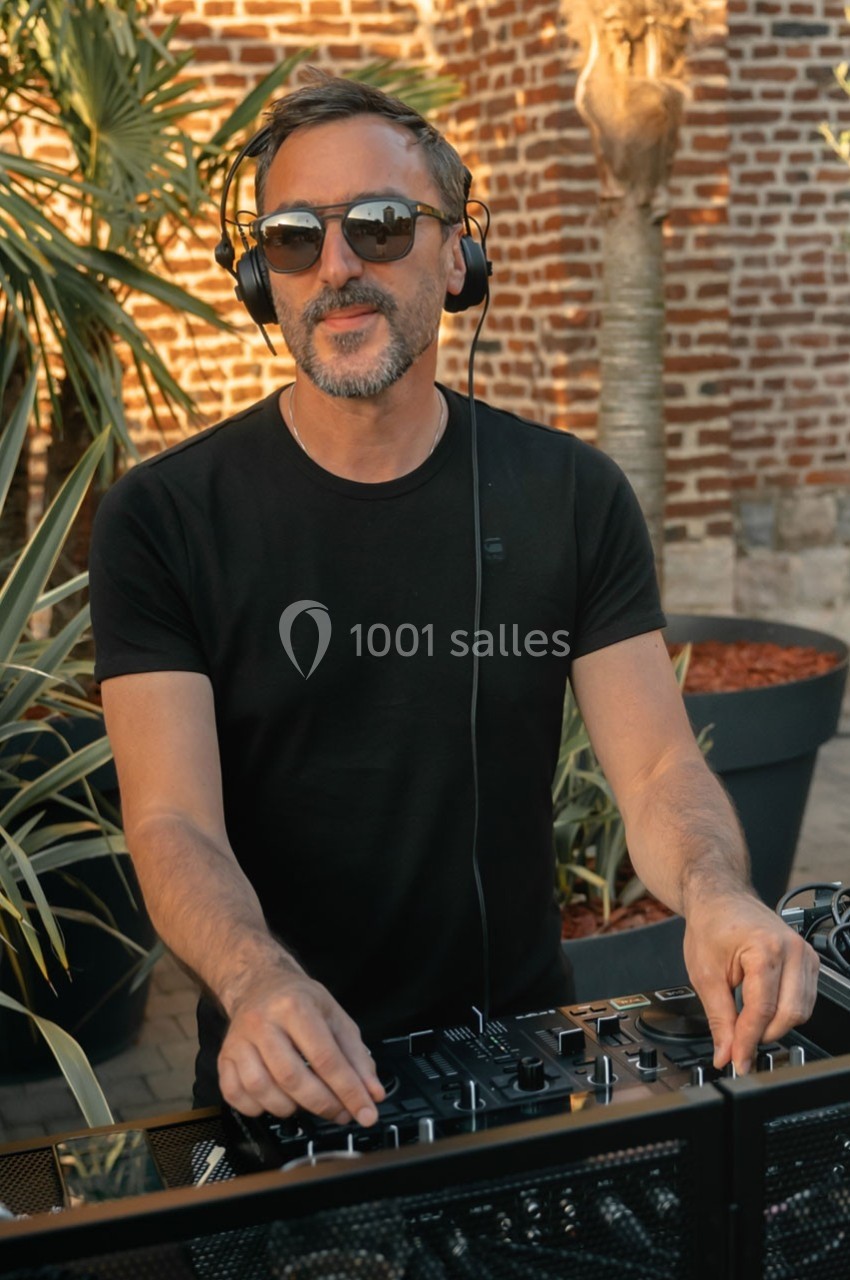 Un DJ mixe en extérieur, portant un casque audio et des lunettes de soleil, avec un mur de briques en arrière-plan.
