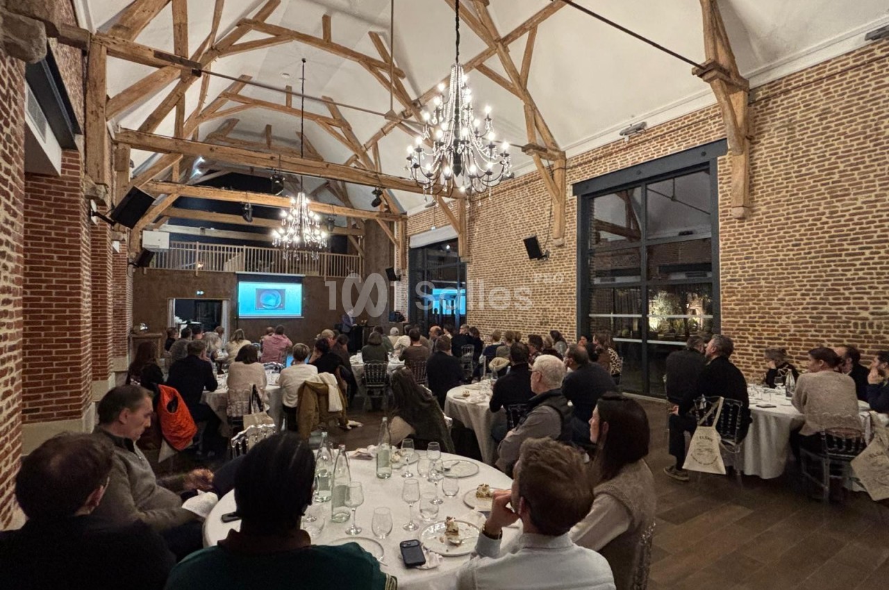 Salle de réception avec des invités assis à des tables, assistant à une présentation projetée sur un écran.