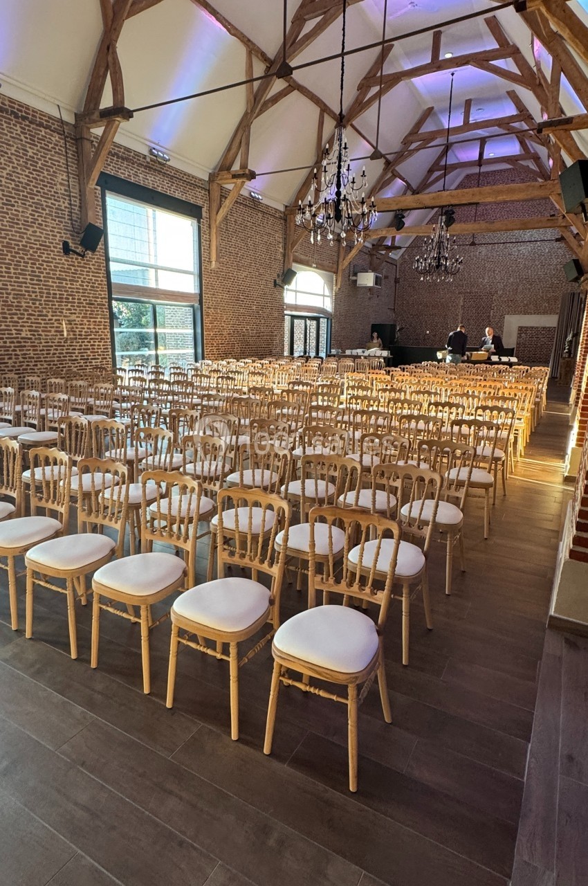 Salle de réception avec rangées de chaises en bois clair et coussins blancs, éclairée par des lustres et des fenêtres.