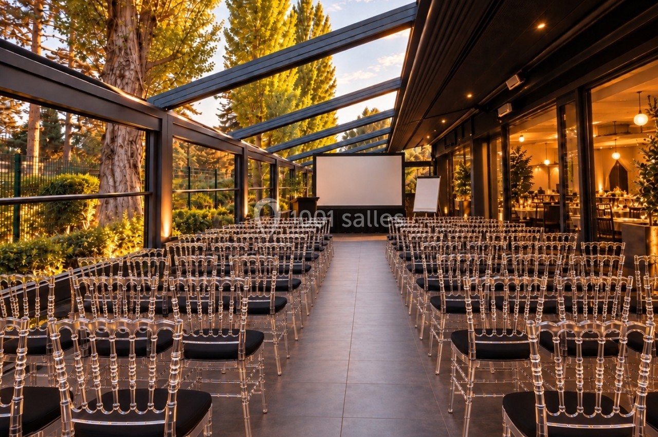 Salle de conférence en plein air avec des chaises transparentes alignées face à un écran de projection.