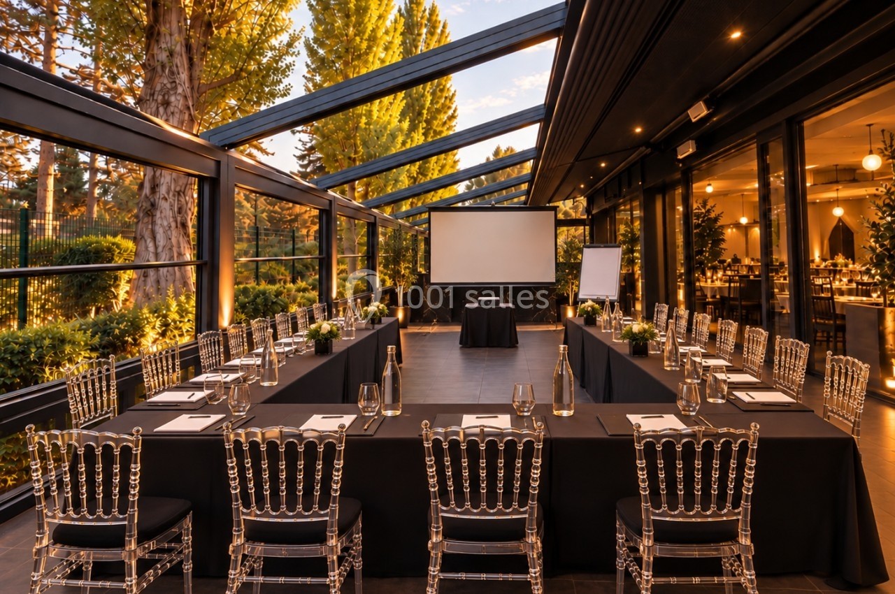 Salle de réunion élégante en extérieur couvert, aménagée en disposition en U avec tables, chaises transparentes et écran.