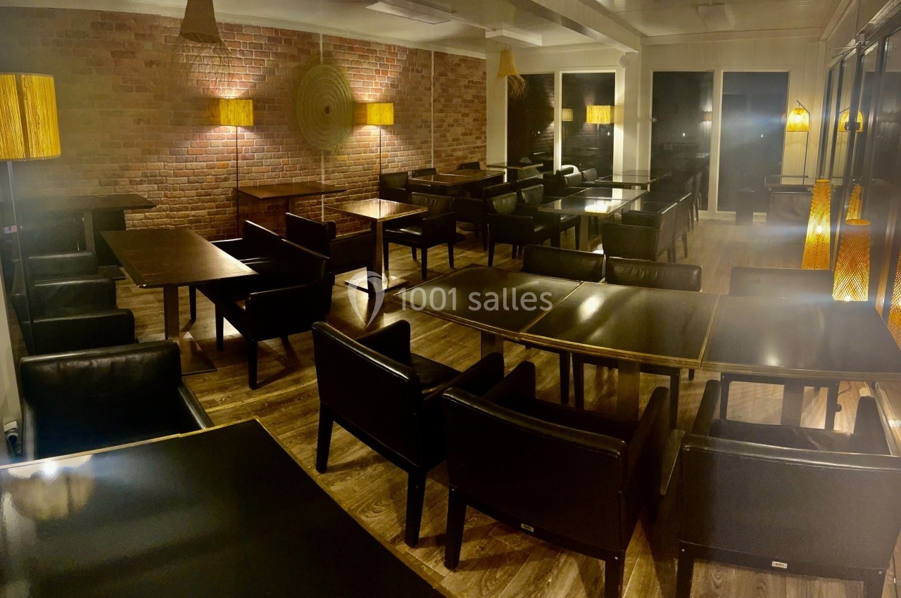 Salle de restaurant vide avec des tables en bois, des fauteuils noirs, un mur en briques et un éclairage tamisé.