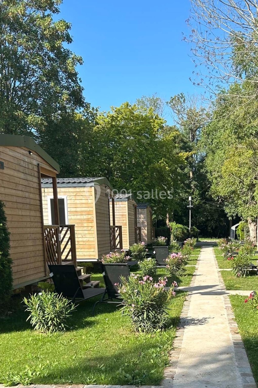 Allée bordée de mobil-homes en bois, entourée de végétation et de fleurs sous un ciel bleu.