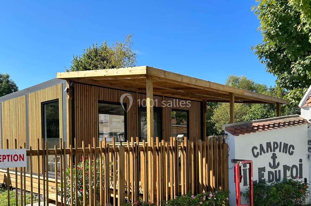 Bâtiment en bois moderne avec terrasse couverte, situé à l'entrée d'un camping, entouré de verdure.