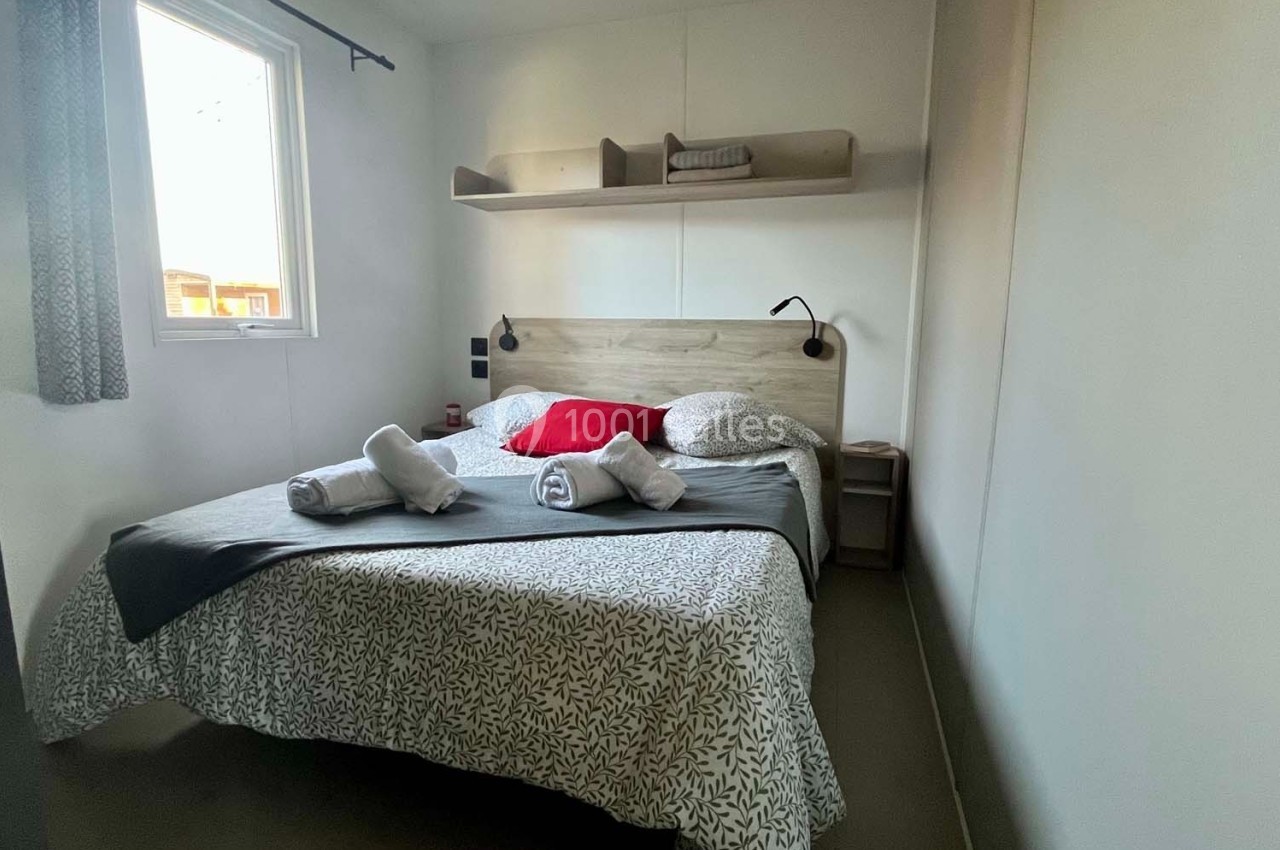 Chambre avec lit double, draps pliés, étagère murale, lampe de chevet et fenêtre laissant entrer la lumière naturelle.
