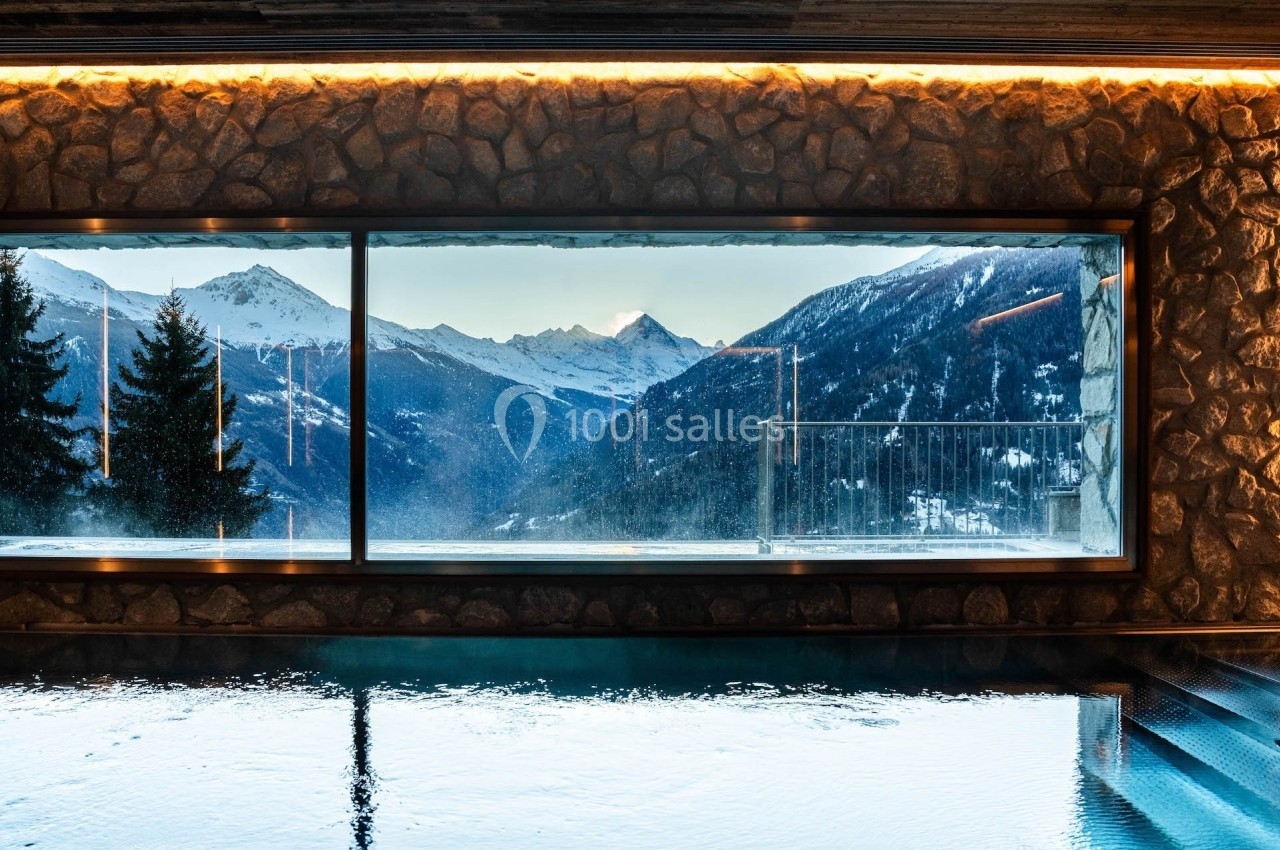 Vue sur des montagnes enneigées à travers une grande baie vitrée depuis une piscine intérieure éclairée.