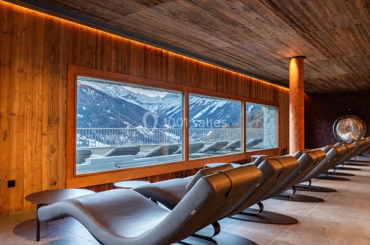 Salle de repos avec chaises longues orientées vers une grande baie vitrée offrant une vue sur des montagnes enneigées.