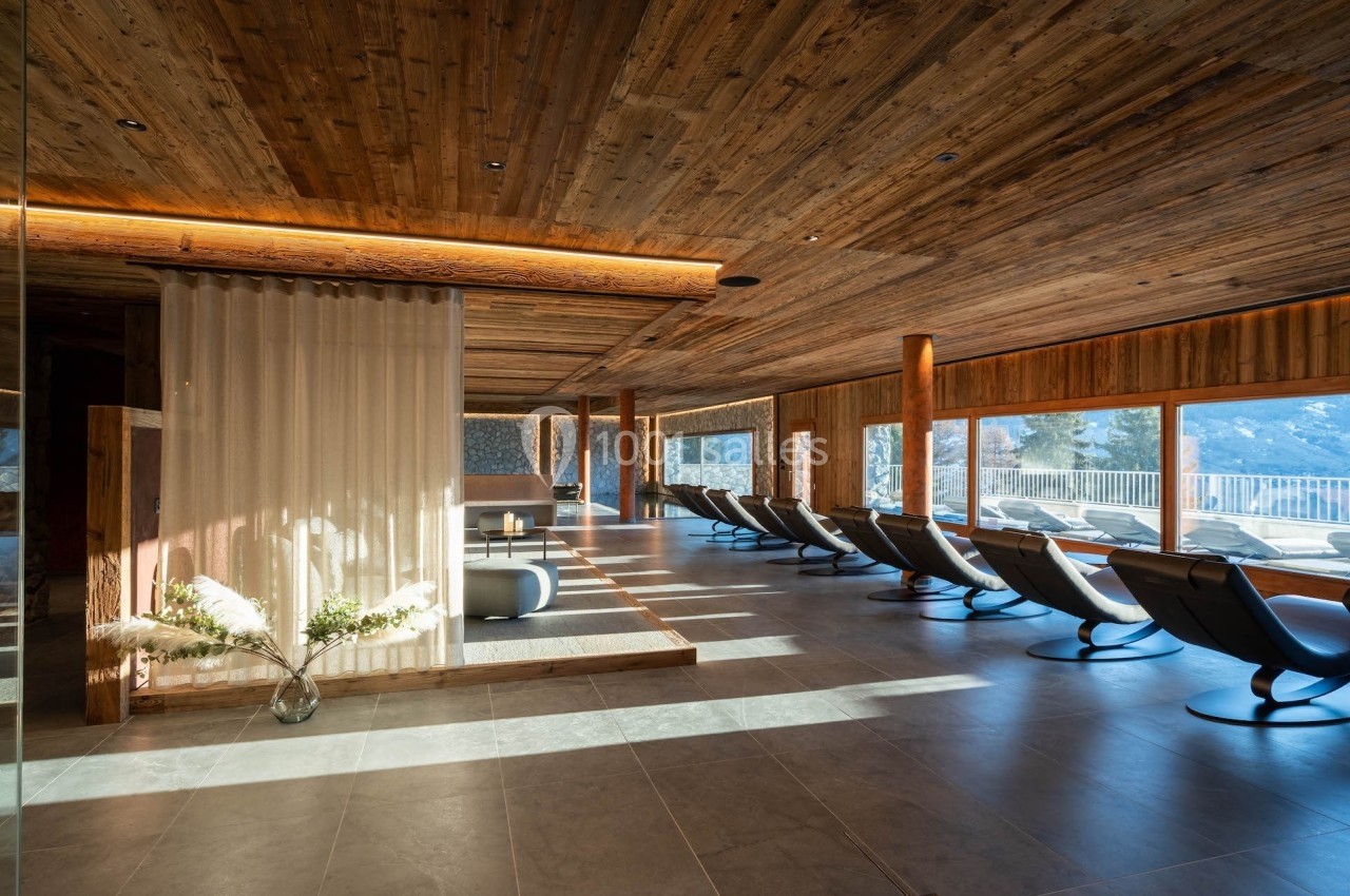 Salle de relaxation lumineuse avec chaises longues, plafond en bois et grandes baies vitrées offrant une vue sur l'extérieur.